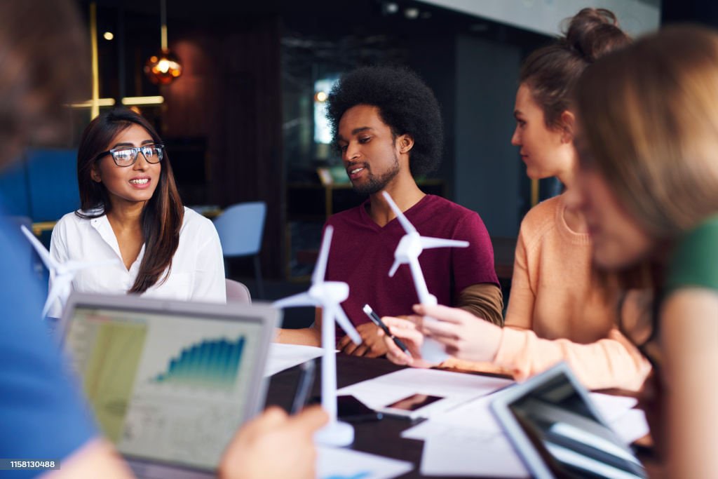 Young professionals discussing offshore wind turbine models during a renewable energy team meeting, with data charts visible on digital devices.